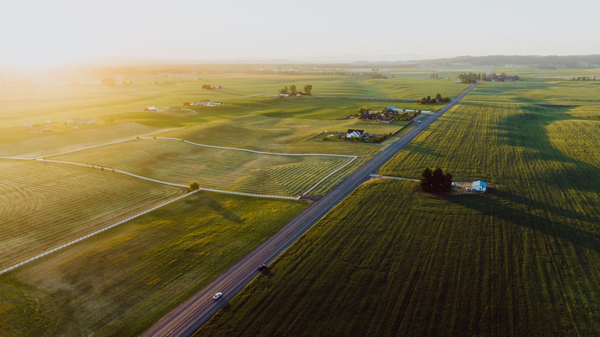 Golden-hour aerial view of open farmland with a long country road and white ranch fencing
